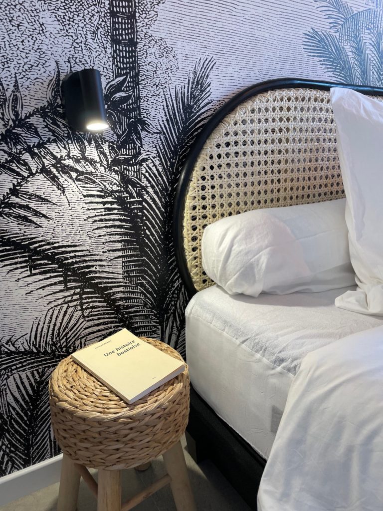 Chambre aux tons naturels avec une tapisserie panoramique représentant des palmiers, une tête de lit en osier agrémentée de touches noires et blanches, créant une ambiance apaisante et contemporaine.