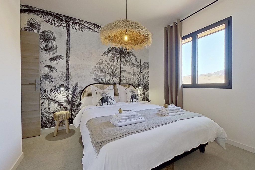 Chambre ethnique élégante avec un lit double 160 cm, décorée d'une tapisserie panoramique représentant des palmiers et un paysage lointain. Suspension en rotin et tonalités apaisantes de beige et noir, créant une ambiance chaleureuse et raffiné.