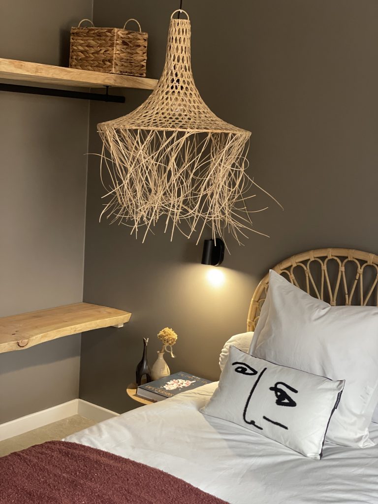 Chambre avec un dressing ouvert composé de tablettes et planches en bois massif brut, agrémentée de suspensions en osier tressé, dans des tons sombres et chaleureux, créant une ambiance naturelle et apaisante.