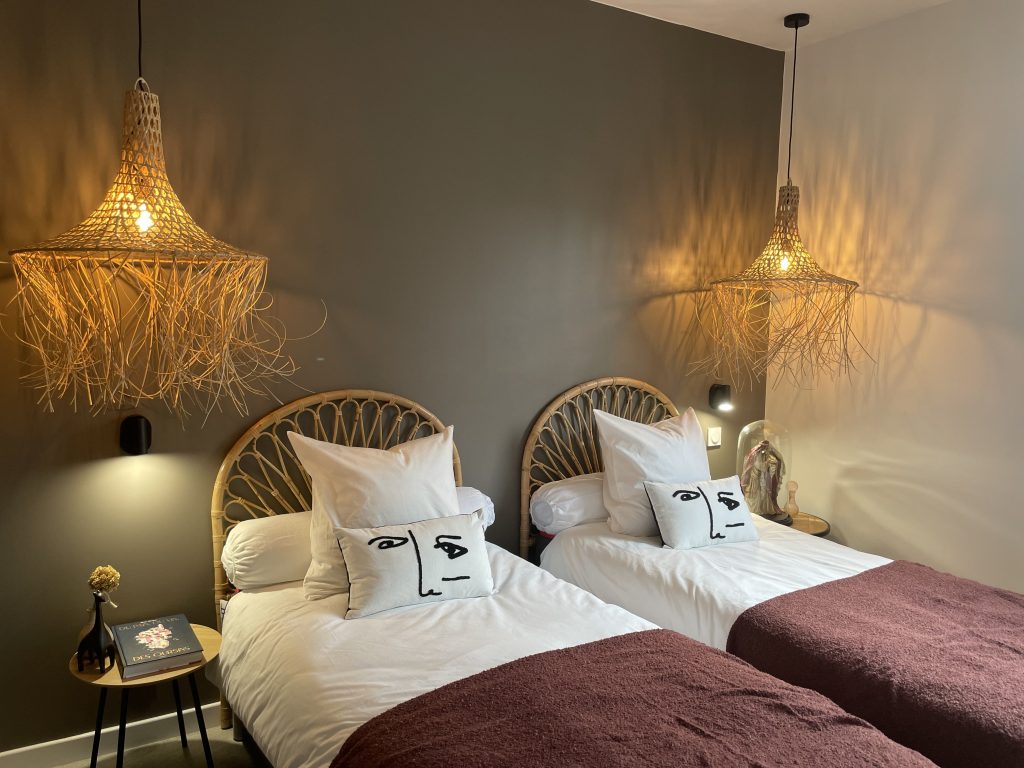 Chambre modulable du projet La Villa Geneviève, avec possibilité d'installer deux lits d'une place ou de les réunir pour créer un grand lit , décorée dans des des tons sombres de taupe, aubergine, gris foncé et beige, avec des suspensions en raffia et têtes de lit en osier tressé, créant une atmosphère tamisée et chaleureuse.