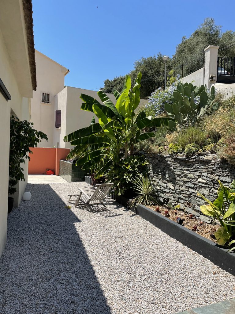 Aménagement extérieur dans un terrain en restanque, avec des murs en pierre sèche, une cuisine d'été en zellige verte rappelant les tonalité kaki de la maison, sol engraviez pour une ambiance méditerranéenne, bananiers et plantes locales ajoutent une touche naturelle et chaleureuse.