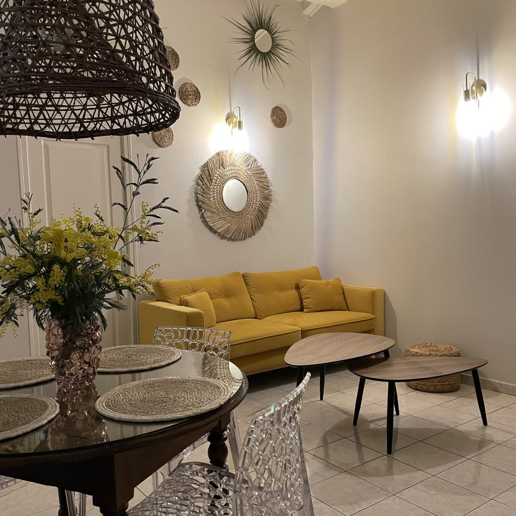 Pièce de vie rénovée avec canapé jaune moutarde, suspension cannage et décoration méditerranéenne, home staging A Casa Cantone en Corse.