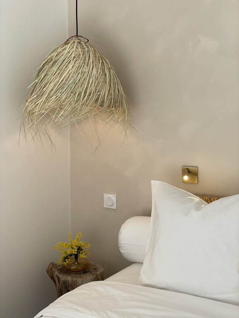 Gros plan sur le lit de la suite parentale, habillé de linge blanc immaculé. En arrière-plan, un mur texturé dans des tonalités chaudes de beige. Une suspension en palmier, une liseuse en laiton doré, des tables de chevets en rondin de bois, créant une ambiance chaleureuse et raffinée.