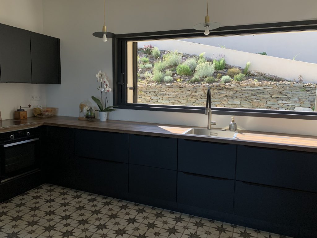 Cuisine contemporaine avec meubles noirs mattes, plans de travail en noyer et crédence en marbre de Sienne mis en valeur par le grand panoramique ouvert sur la nature environnante.