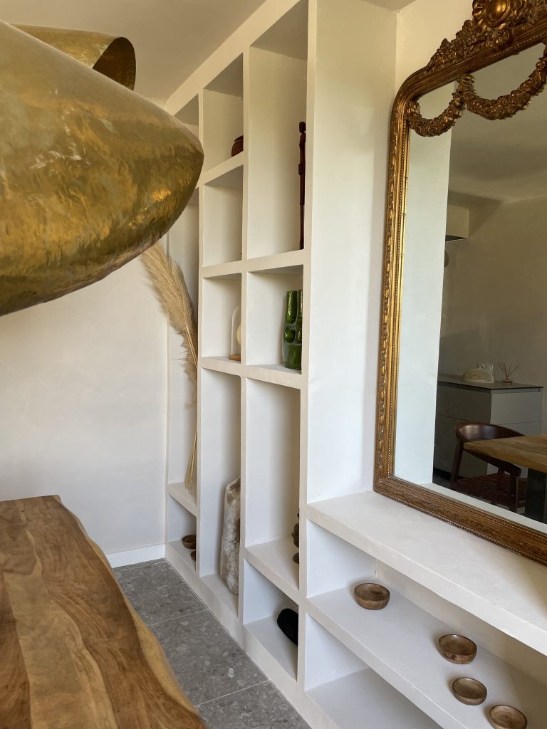Bibliothèque murale à la chaux avec niches décoratives, objets d'art, vases, plantes vertes, miroir doré ancien et sol en pierre grise, table en bois, luminaires en laiton doré, créant une ambiance élégante et épurée.