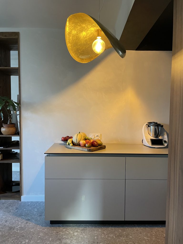 Cuisine moderne avec plan de travail beige, meubles minimalistes sans poignées, robots de cuisine, fruits frais, sol en pierre grise, tableau artistique de bœuf, plantes vertes et luminaire en laiton doré