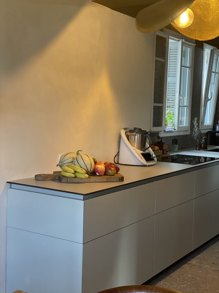 Cuisine moderne avec plan de travail beige, meubles minimalistes sans poignées, robots de cuisine, fruits frais, sol en pierre grise, et luminaire en laiton doré