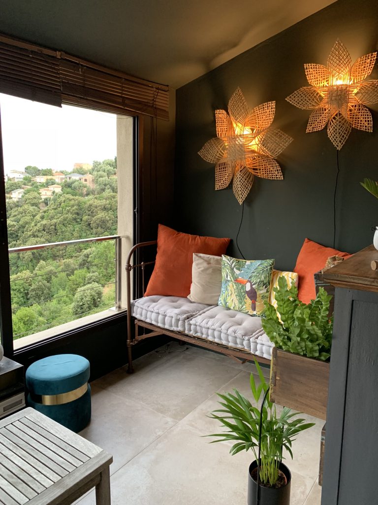 Véranda aménagée comme un cocon suspendu au bord de la vallée, telle un nid d'aigle. Ambiance chaleureuse avec des appliques représentant de grandes fleurs en raphia et des touches de orange, vert bleu.