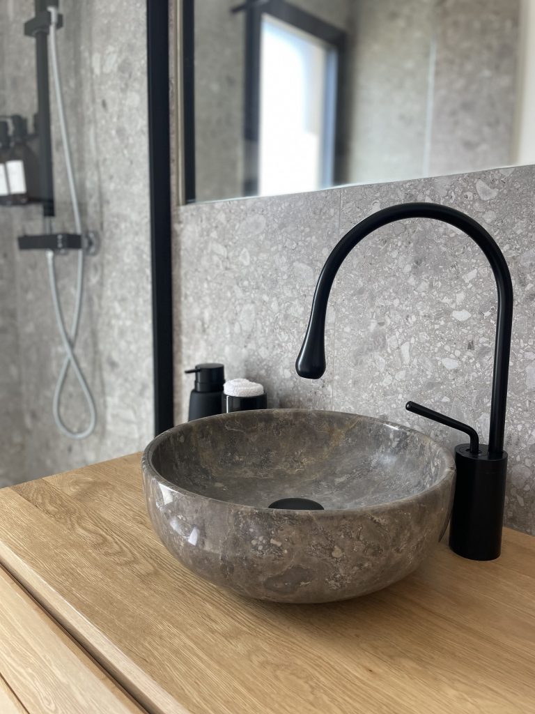 Salle d'eau de la suite parentale avec un meuble vasque en chêne massif, vasque en marbre grège, carrelage imitation pierre dans des tons minéraux, robinetterie et accessoires noirs, créant un contraste élégant entre matière noble et touches contemporaines.