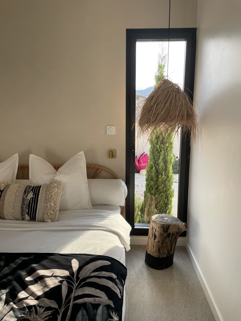 Chambre de la suite parentale du projet La Villa Genviève, avec grand lit king size, décoration chaleureuse, chevet en tronc d'arbre, suspensions en palmiers et châssis vitré offrant une vue apaisante sur la nature environnante.