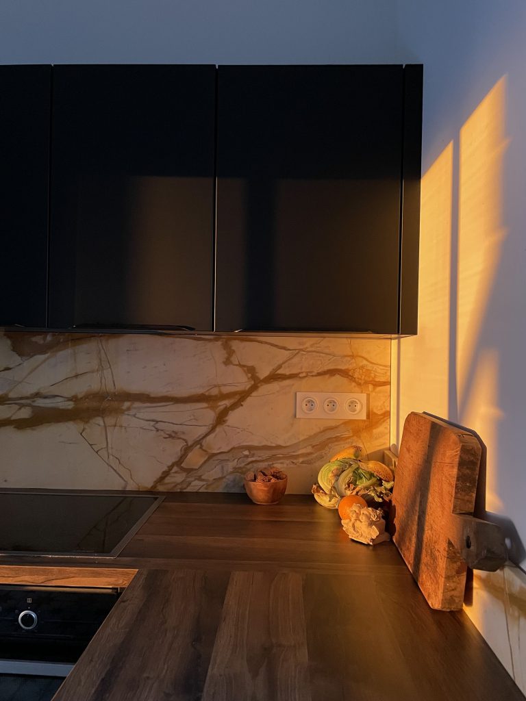 Cuisine contemporaine du projet Pietrabugno avec des meubles noirs mattes, plan de travail en noyer et crédence en marbre de Sienne aux tonalités orange, jaune et brun, créant un contraste entre modernité et tradition.