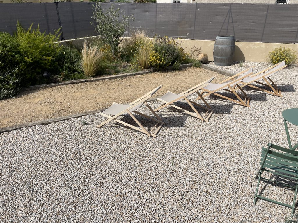Jardin de l'entrée du projet La Villa Geneviève avec terrain de pétanque en tuf damé, enfilade de chiliennes pour se détendre en regardant les amis jouer, sol recouvert de gravier, créant un espace convivial et chaleureux pour profiter de l'extérieur.