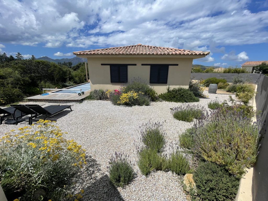 Vue d'ensemble de La Villa Geneviève, villa rectangulaire avec une toiture 4 pentes de style méditerranéen avec des lignes contemporaines, menuiseries élégantes et entourée d'une végétation typique de la Corse au abord de la piscine, près de Calvi.