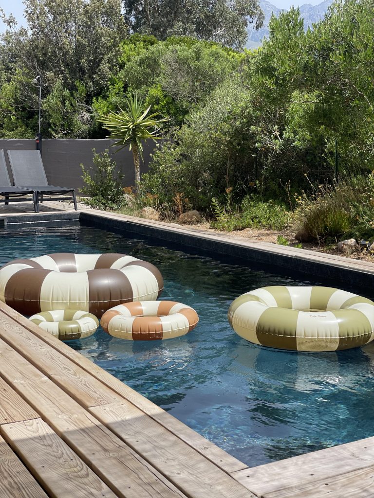 Terrasse de La Villa Geneviève avec une grande piscine couleur rivière, ensemble de bouées de la marque Petite Pomme, transats confortables pour la détente, le tout entouré de verdure et du maquis corse.