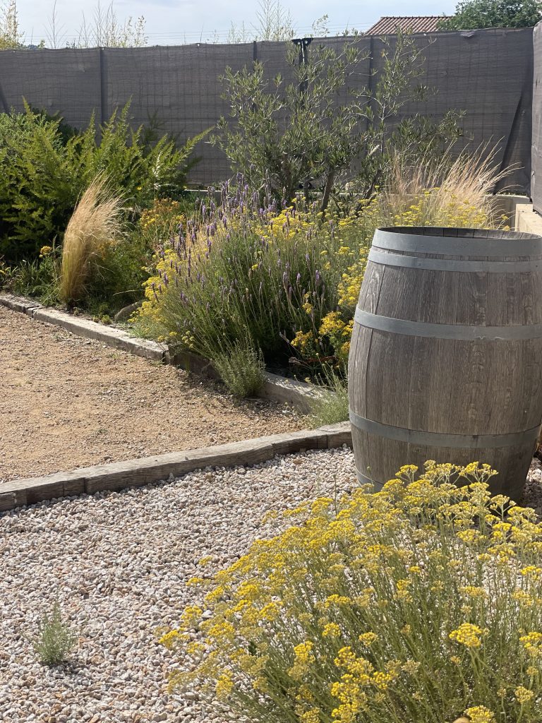 Terrain de pétanque entouré d'immortelles, de romarins, de cheveux d'anges et d'oliviers, avec un tonneau de vignoble, offrant un extérieur authentique et méditerranéen.