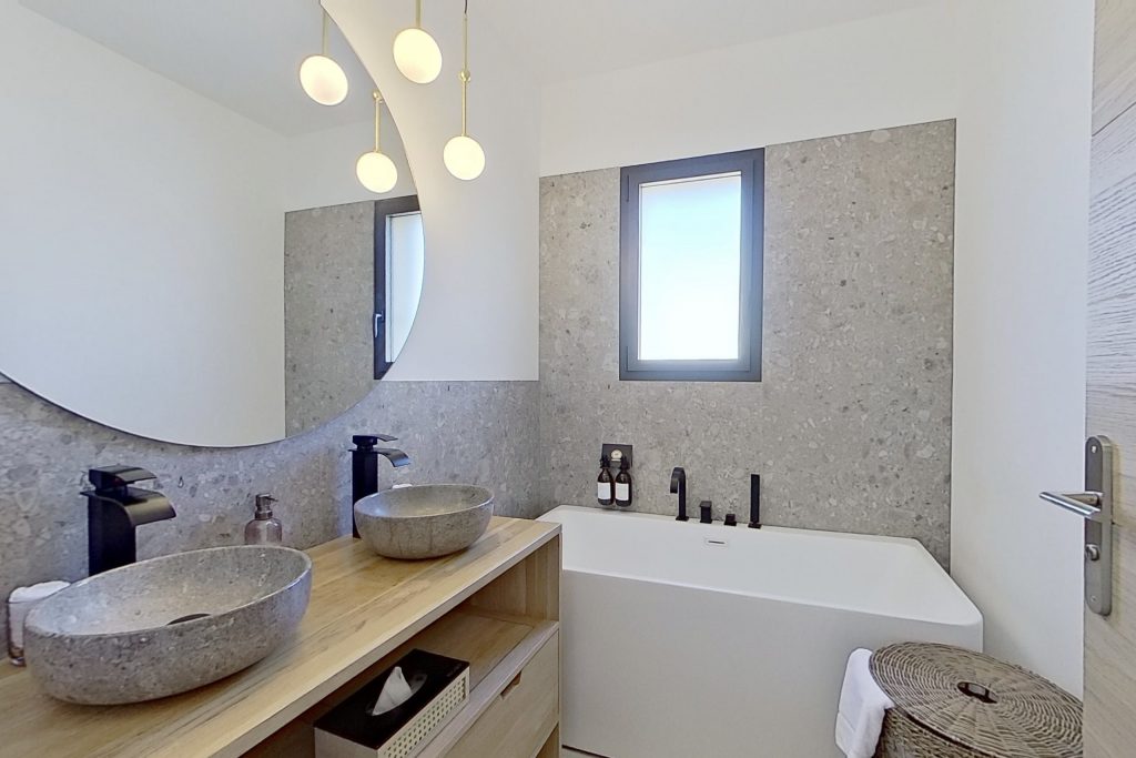 Salle de bain élégante avec des meubles en chêne, double vasque en marbre, baignoire semi îlot blanche. Le sol et les murs sont carrelés en grès cérame imitation terrazzo, complétés par des accessoires de robinetterie noirs pour un contraste moderne.