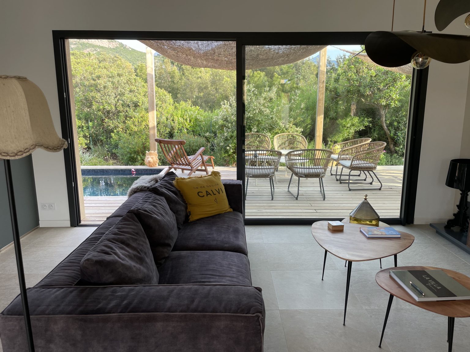 Salon du projet La Villa Geneviève avec vue sur la terrasse, la végétation méditerranéenne, et une partie de la piscine, créant une atmosphère ouverte et lumineuse dans un cadre naturel et apaisant.