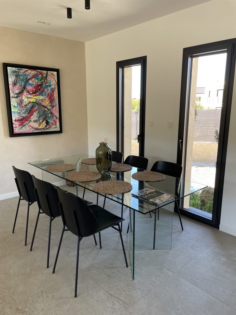 Salle à manger avec une grande table en verre pouvant accueillir 8 personnes, mise en valeur par un tableau de l'artiste peintre Alain Soma exposé au mur, dans une ambiance élégante et épurée.