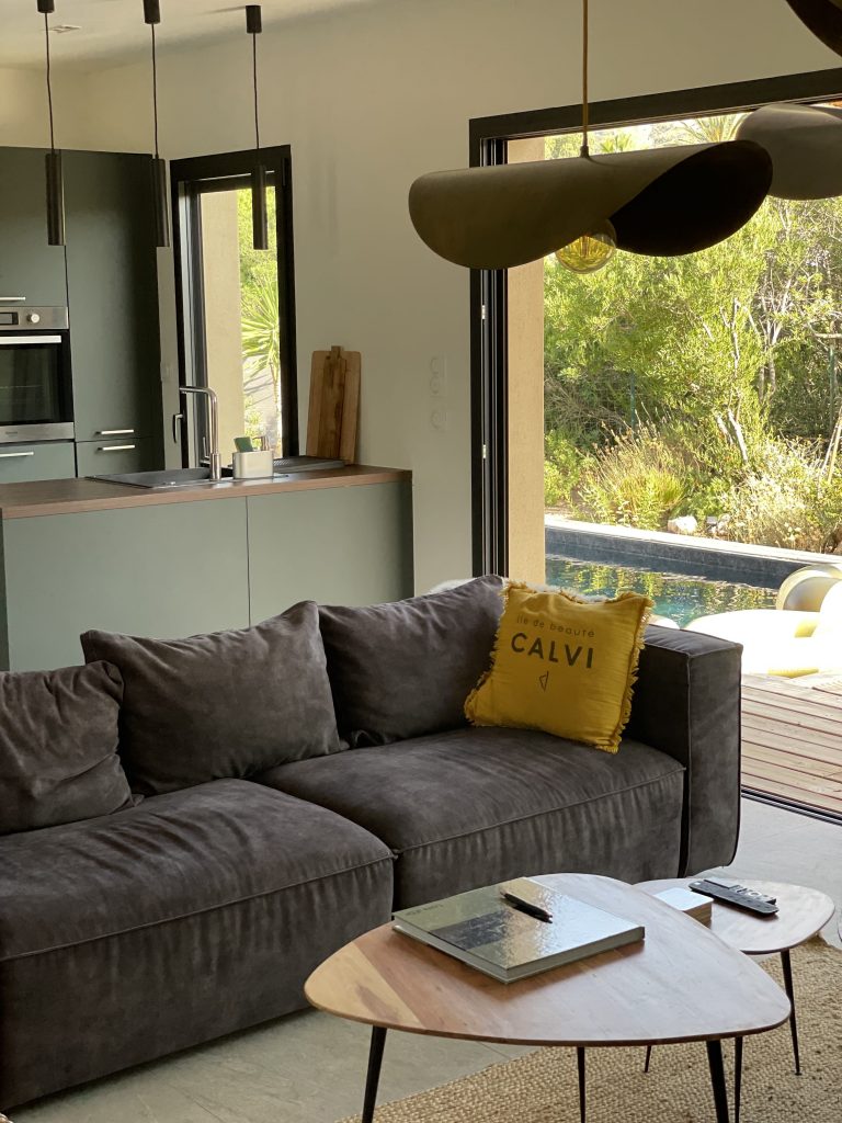 Vue depuis le salon du projet La Villa Geneviève donnant accès à cuisine ouverte aux tons kaki, avec accès direct à la terrasse et à la piscine couleur rivière, créant une harmonie entre les espaces intérieurs et extérieurs dans un cadre naturel.