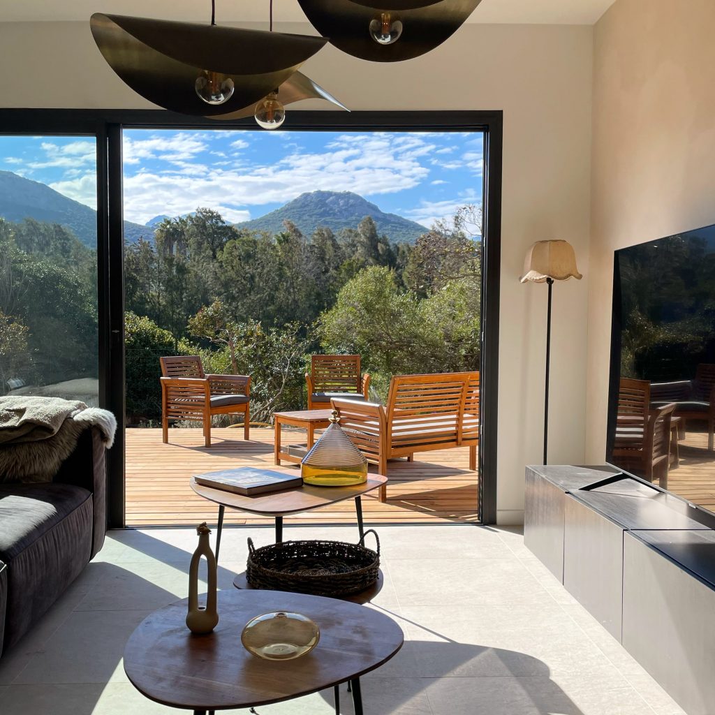 Salon lumineux du projet La Villa Geneviève avec baie de 4 mètres offrant une vue panoramique sur le paysage de la balagne et les montagnes corses, mobilier en bois, suspensions aériennes, tons naturels et chaleureux, terrasse en bois prolongeant l'espace vers l'extérieur