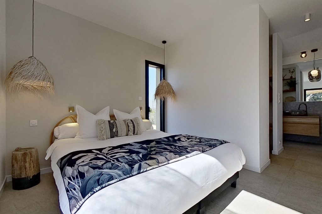 Chambre de la suite parentale avec un grand lit, baignée de lumière, aux tons naturels et chaleureux. Mur à la chaux, suspensions en fibres de palmiers et rondins de bois en guise de tables de chevet, avec une perspective ouverte sur le dressing et la salle de bain attenante, créant une atmosphère douce et harmonieuse.