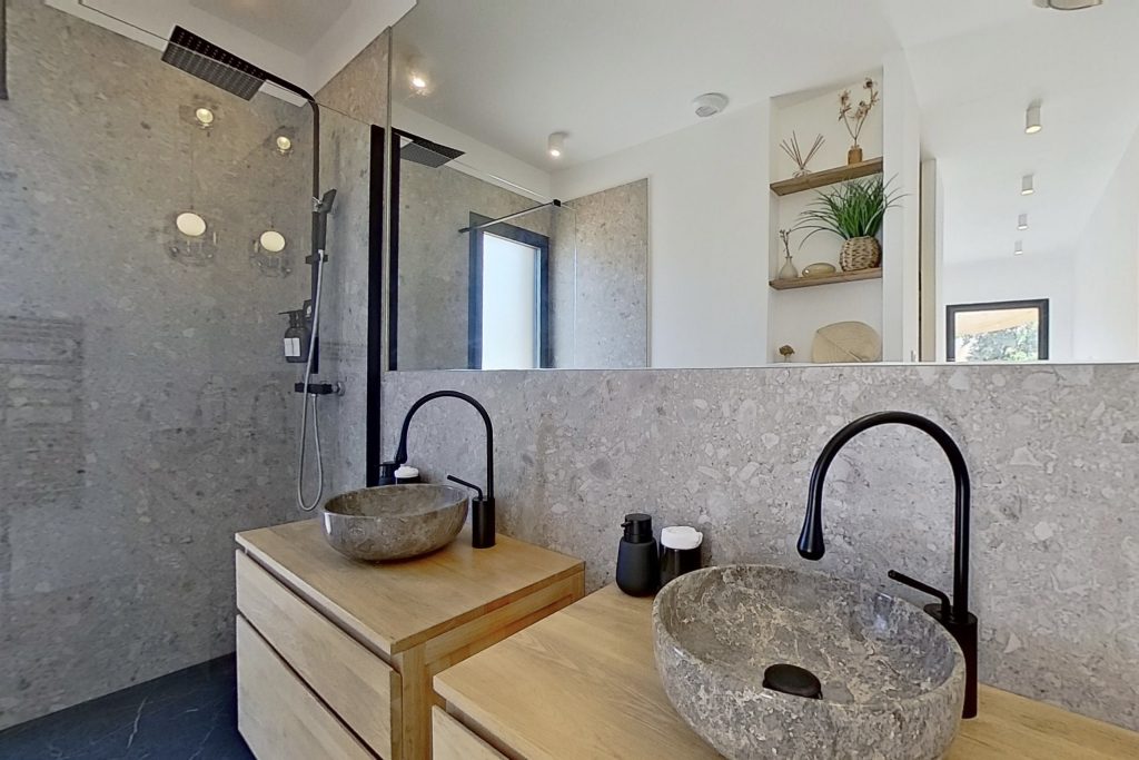 Salle d'eau de la suite parentale avec deux meubles vasques en chêne massif, vasque en marbre grège, carrelage imitation pierre dans des tons minéraux, robinetterie et accessoires noirs, créant un contraste élégant entre matière noble et touches contemporaines.