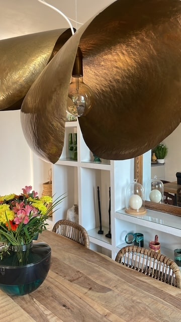 Bibliothèque murale à la chaux avec niches décoratives, objets d'art, vases, plantes vertes, miroir doré ancien, suspension en laiton doré, table en bois massif, créant une ambiance élégante et épurée.