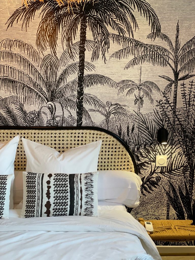 Troisième chambre du projet La Villa Geneviève avec grand lit double, décorée de motifs méditerranéens et africains, tapisserie panoramique en noir et blanc représentant un paysage de palmiers, et éléments en paille et osier tressé, créant une ambiance exotique et élégante.